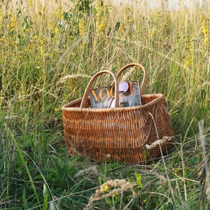 May include: A brown wicker basket with two handles sits in a field of tall green grass. The basket contains a magazine with the word "Vogue" on the cover.
