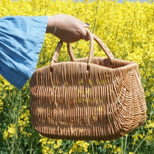May include: A woven wicker basket with two handles, held by a hand in a blue shirt, against a backdrop of yellow flowers.