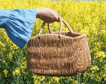 May include: A woven wicker basket with two handles, held by a hand in a blue shirt, against a backdrop of yellow flowers.