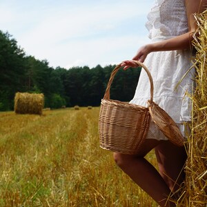 May include: A woman in a white dress holds a wicker basket in a field of hay bales.