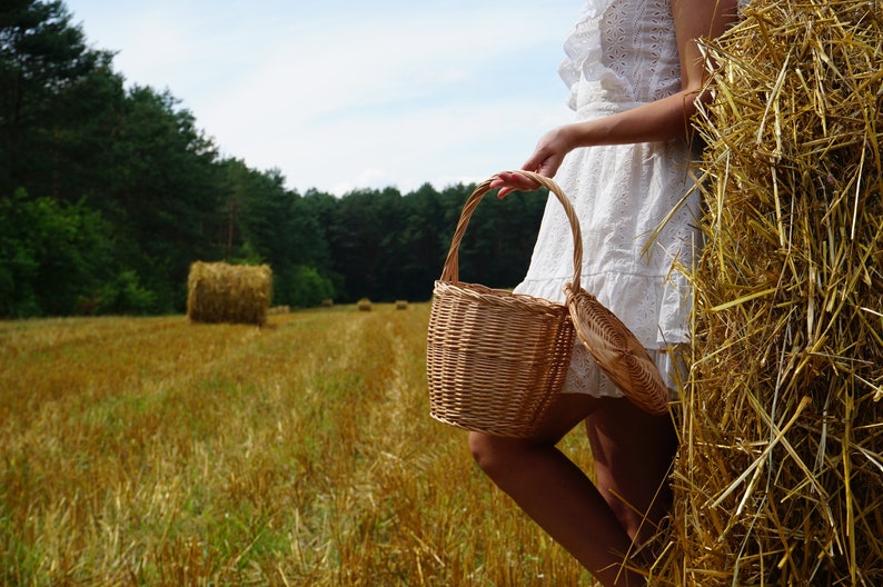May include: A woman in a white dress holds a wicker basket in a field of hay bales.