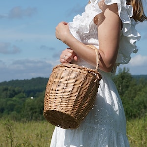May include: A woman wearing a white dress holds a woven wicker basket with a handle. The basket is brown and has a natural woven texture. The woman is standing in a field with green grass and trees in the background.