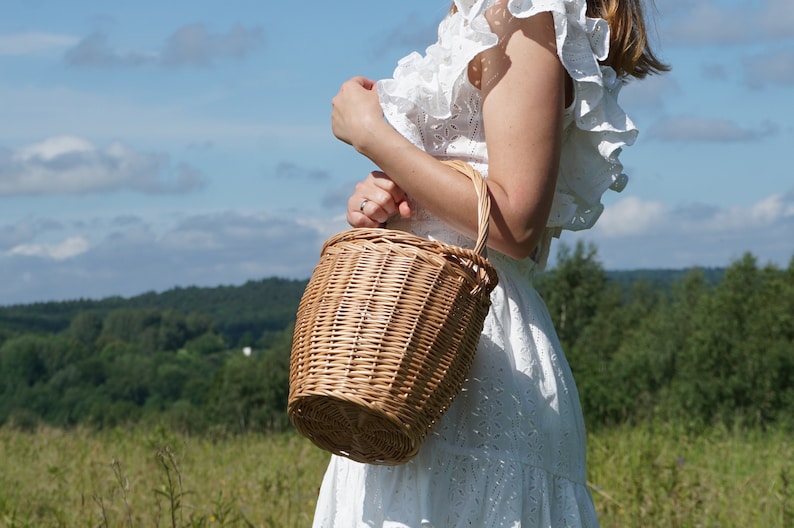 May include: A woman wearing a white dress holds a woven wicker basket with a handle. The basket is brown and has a natural woven texture. The woman is standing in a field with green grass and trees in the background.