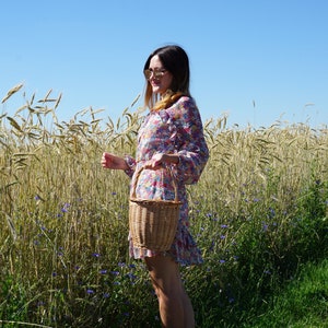 May include: A woman in a floral dress and sunglasses stands in a field of wheat, holding a wicker basket.