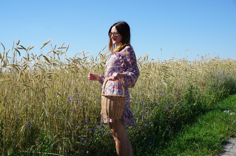 May include: A woman in a floral dress and sunglasses stands in a field of wheat, holding a wicker basket.