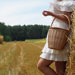 May include: A woman in a white dress with a ruffled hem stands in a field of hay bales. She is holding a wicker basket with a handle.