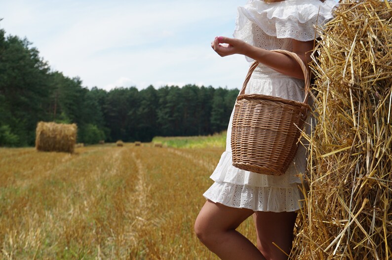 May include: A woman in a white dress with a ruffled hem stands in a field of hay bales. She is holding a wicker basket with a handle.