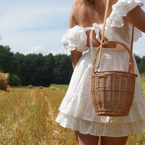 May include: A woman in a white dress with a wicker basket bag with a leather strap. The woman is standing in a field of hay bales.