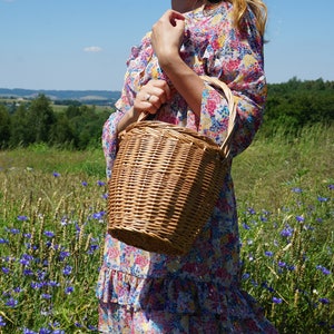 May include: A woman in a floral dress holds a woven wicker basket with a handle. The basket is brown and has a natural woven texture. The woman is standing in a field of wildflowers.