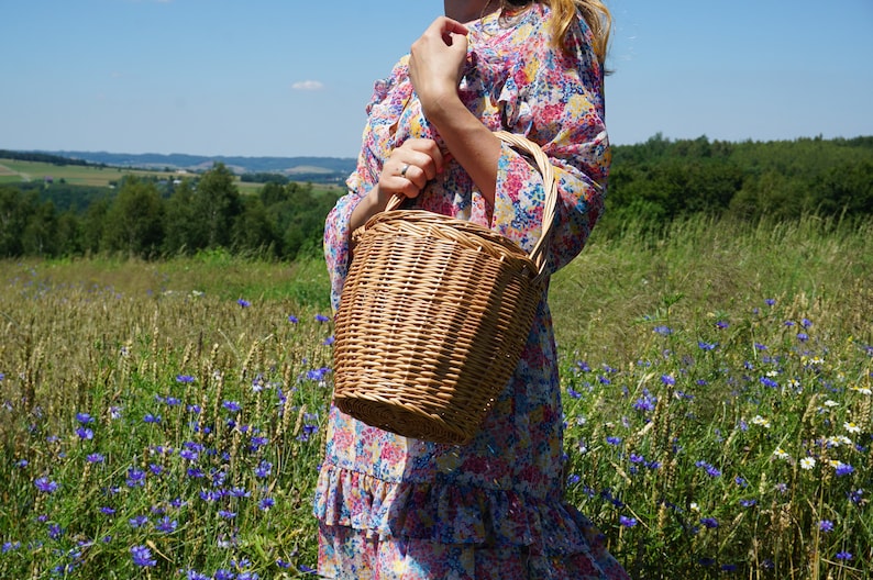 May include: A woman in a floral dress holds a woven wicker basket with a handle. The basket is brown and has a natural woven texture. The woman is standing in a field of wildflowers.