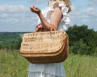 May include: A woman in a white dress holds a large, woven wicker basket with a brown handle. The basket is in focus, and the woman and the background are blurred.