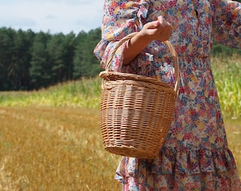 May include: A woman in a floral dress carries a woven wicker basket with a handle. The basket is brown and has a natural woven texture. The woman is standing in a field of tall grass.