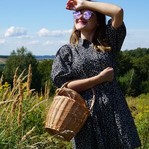 May include: A woman wearing a black and white floral dress and sunglasses holds a wicker basket in a field.