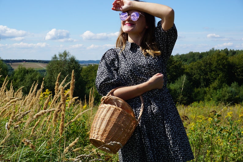 May include: A woman wearing a black and white floral dress and sunglasses holds a wicker basket in a field.