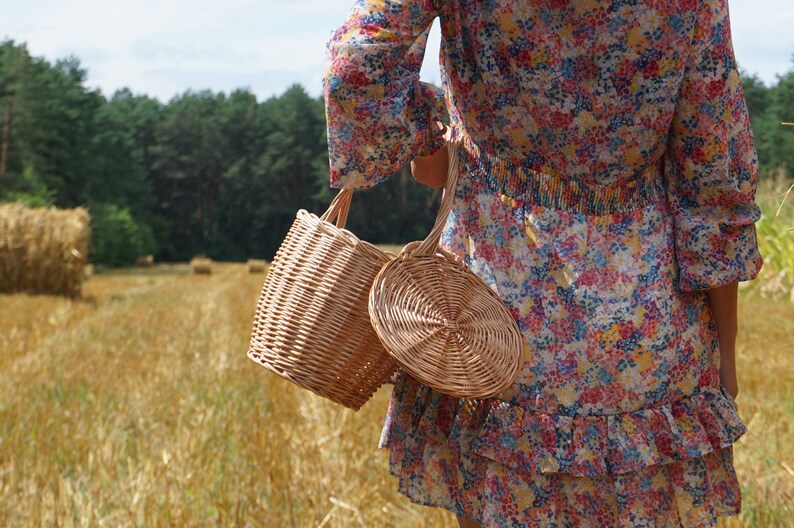 May include: Two wicker baskets, one round and one cylindrical, are held by a person wearing a floral dress. The baskets are made of natural woven material and have a rustic, handmade look.
