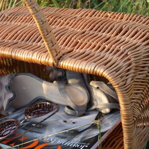 May include: A brown wicker basket with a handle, containing a pair of sandals and a magazine. The basket is sitting on a bed of green grass.