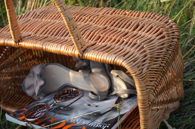 May include: A brown wicker basket with a handle, containing a pair of sandals and a magazine. The basket is sitting on a bed of green grass.