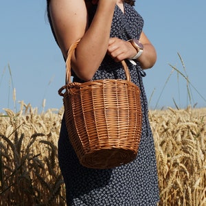 May include: A brown wicker basket bag with a handle. The bag is being held by a person in a field of wheat.