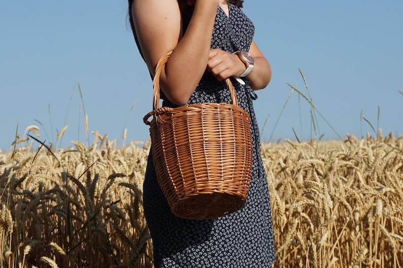 May include: A brown wicker basket bag with a handle. The bag is being held by a person in a field of wheat.