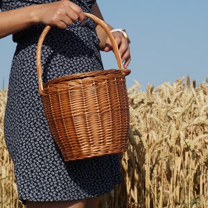 May include: A woman in a blue floral dress holds a brown wicker basket with a handle in a field of wheat.