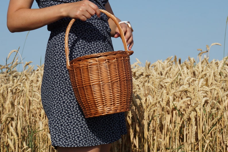 May include: A woman in a blue floral dress holds a brown wicker basket with a handle in a field of wheat.