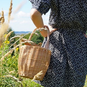 May include: A woman in a black and white floral dress holds a woven wicker basket with a handle.