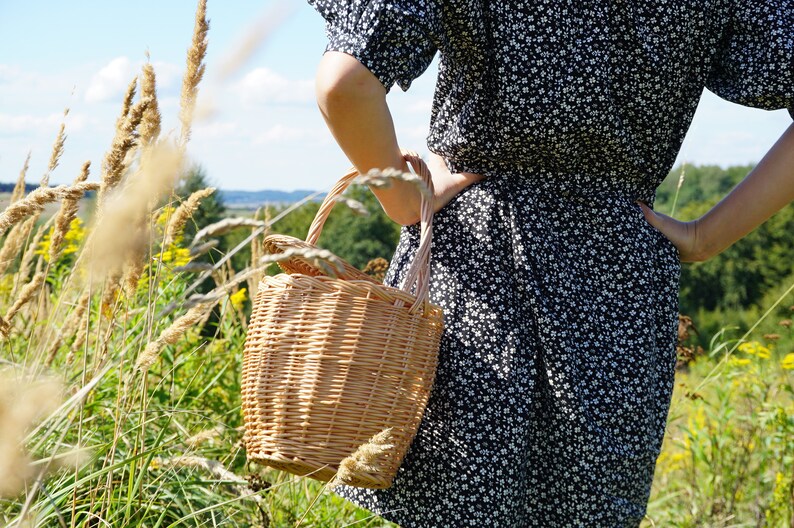 May include: A woman in a black and white floral dress holds a woven wicker basket with a handle.