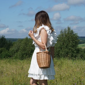 May include: A woman wearing a white dress with ruffles stands in a field holding a wicker basket bag.