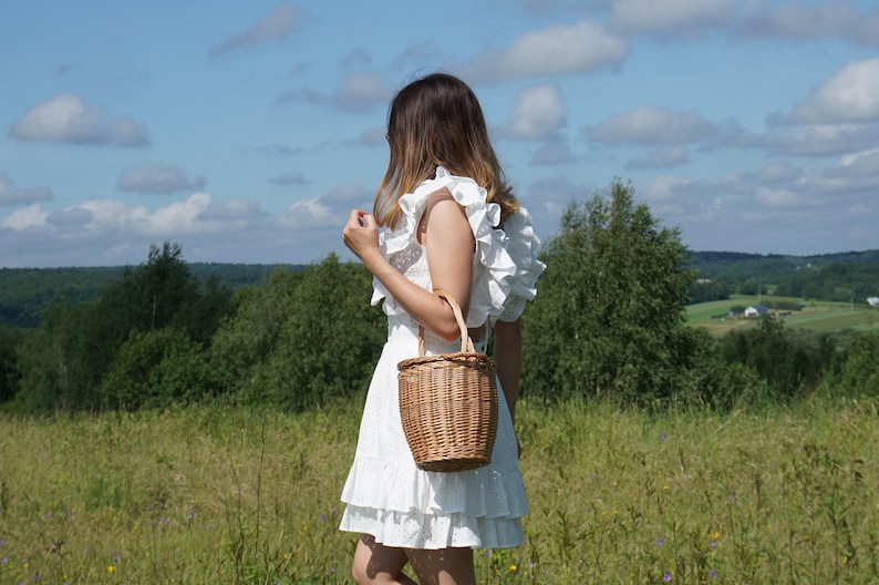 May include: A woman wearing a white dress with ruffles stands in a field holding a wicker basket bag.