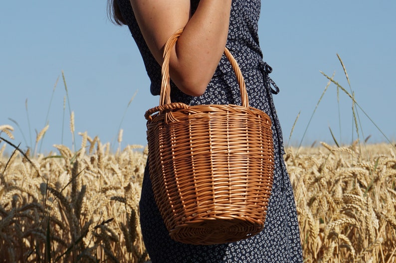 May include: A brown wicker basket with a woven handle. The basket is round and has a slightly flared top. It is being held by a person in a field of wheat.