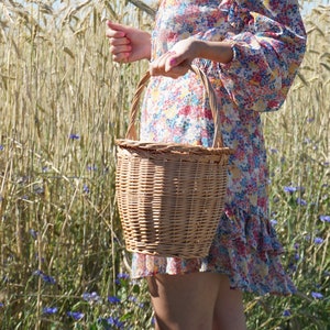 May include: A woman in a floral dress holds a woven wicker basket with a handle in a field of wheat.