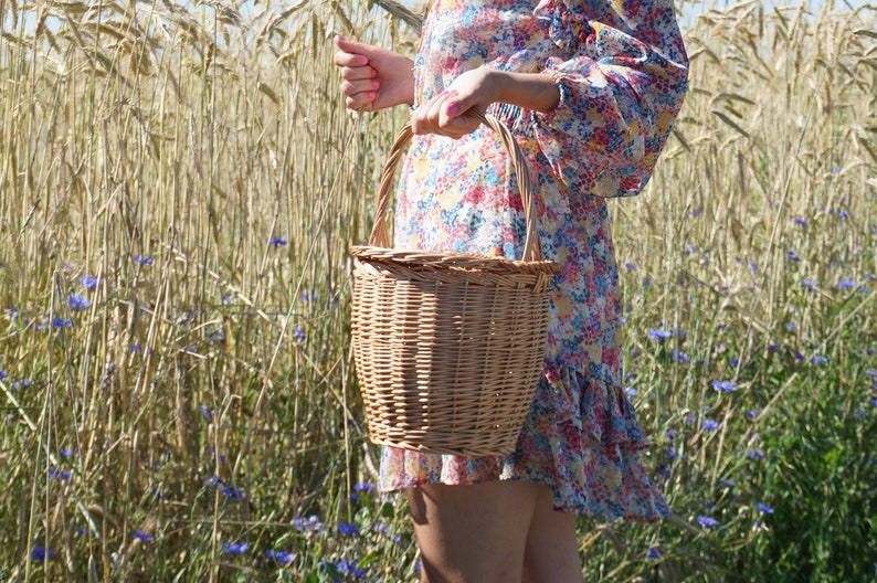 May include: A woman in a floral dress holds a woven wicker basket with a handle in a field of wheat.