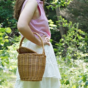 May include: A woman in a pink sleeveless shirt and white skirt holds a wicker picnic basket with a handle.