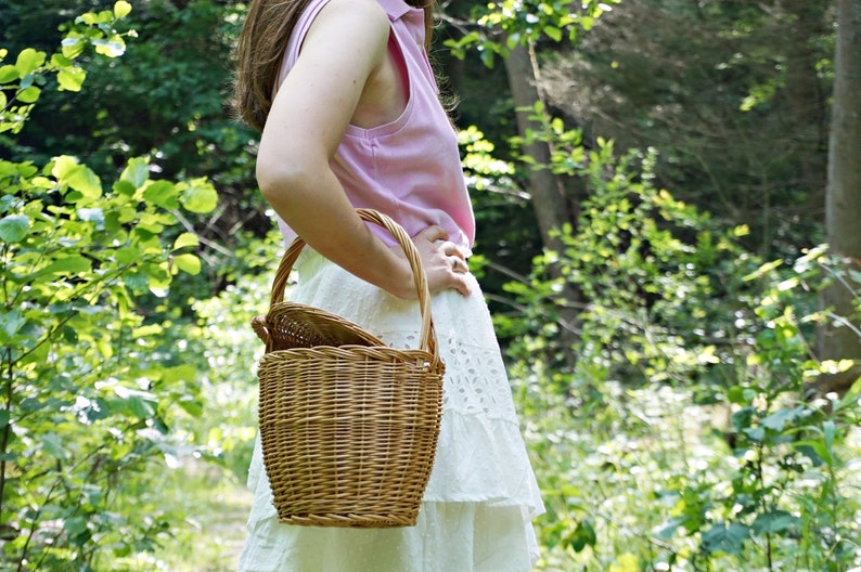 May include: A woman in a pink sleeveless shirt and white skirt holds a wicker picnic basket with a handle.