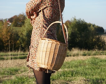 May include: A woven wicker basket with a leather strap and handle. The basket is being worn as a crossbody bag.