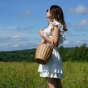 May include: A woman wearing a white dress with frills stands in a field holding a wicker basket. The sky is blue with white clouds.