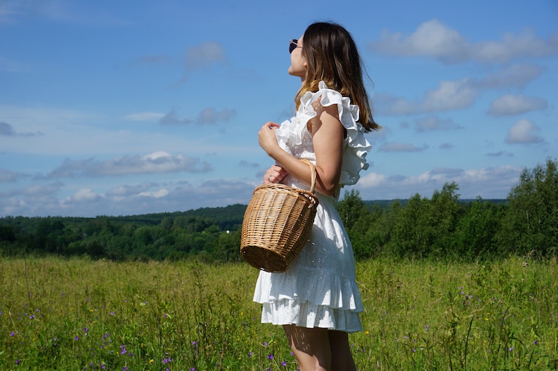 May include: A woman wearing a white dress with frills stands in a field holding a wicker basket. The sky is blue with white clouds.