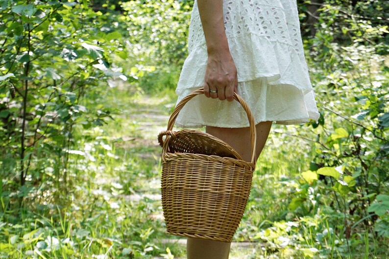 May include: A woman in a white dress carries a woven wicker basket with a handle in a forest setting.
