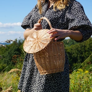 May include: A woven wicker basket with a round lid. The basket is light brown and has a natural finish. The basket is being held by a person in a field.