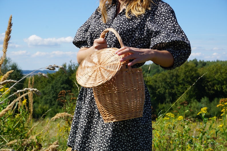 May include: A woven wicker basket with a round lid. The basket is light brown and has a natural finish. The basket is being held by a person in a field.