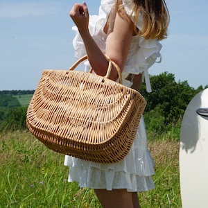 May include: A woman in a white dress holds a woven wicker basket with a handle. The basket is large and has a rounded shape. The woman is standing in a field of green grass with a white car in the background.