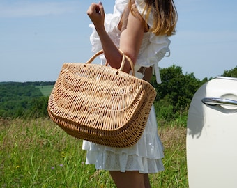 May include: A woman wearing a white dress holds a woven wicker basket with a handle. The basket is large and has a rounded shape. The woman is standing in a field of green grass with a white car in the background.