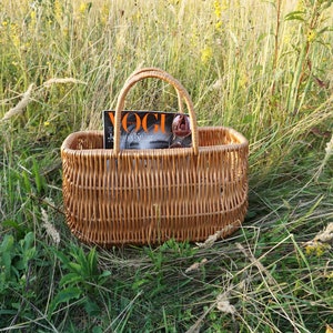 May include: A woven wicker basket with a handle, sitting in a field of tall grass. A magazine with the word "Vogue" on the cover is inside the basket.