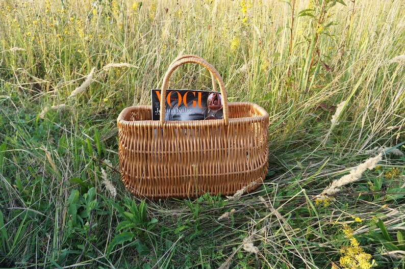 May include: A woven wicker basket with a handle, sitting in a field of tall grass. A magazine with the word "Vogue" on the cover is inside the basket.