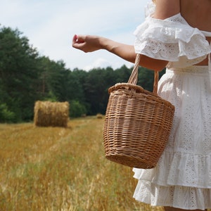May include: A woman in a white eyelet dress with a ruffled hem holds a woven wicker basket bag in a field of hay bales.