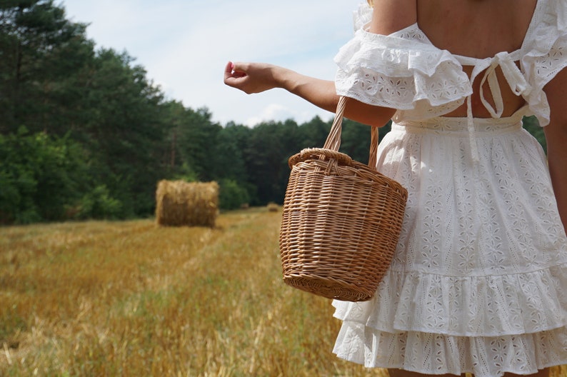 May include: A woman in a white eyelet dress with a ruffled hem holds a woven wicker basket bag in a field of hay bales.