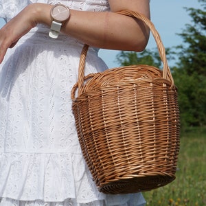 May include: A woman in a white dress with a wicker basket bag. The basket is brown and has a woven design. The woman is wearing a watch on her wrist.