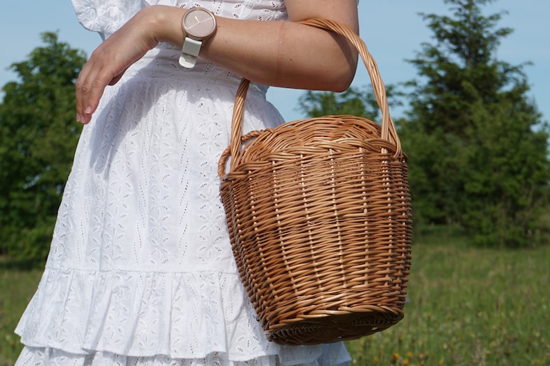 May include: A woman in a white dress with a wicker basket bag. The basket is brown and has a woven design. The woman is wearing a watch on her wrist.