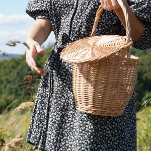 May include: A wicker basket with a lid, held by a person wearing a black and white floral dress. The basket is made of natural woven material and has a round shape.