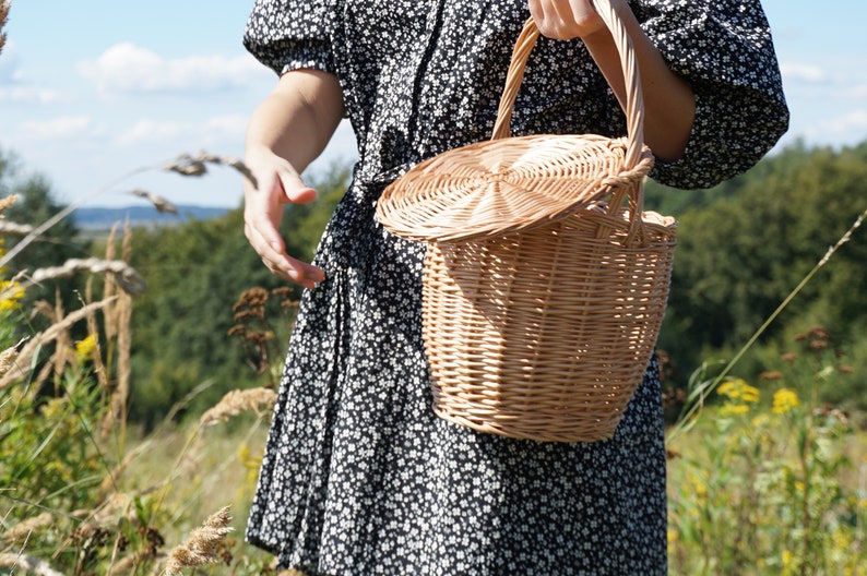 May include: A wicker basket with a lid, held by a person wearing a black and white floral dress. The basket is made of natural woven material and has a round shape.